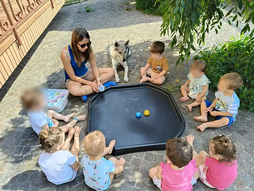 Un gruppo di bambini con la maestra stanno giocando all'esterno della struttura. Assieme a loro uno dei due cani che accompagna le loro avventure. 